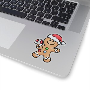 May include: A sticker featuring a cartoon gingerbread man wearing a Santa hat and holding a candy cane. The gingerbread man has a smiling face and is decorated with colorful buttons. The sticker is placed on a laptop.