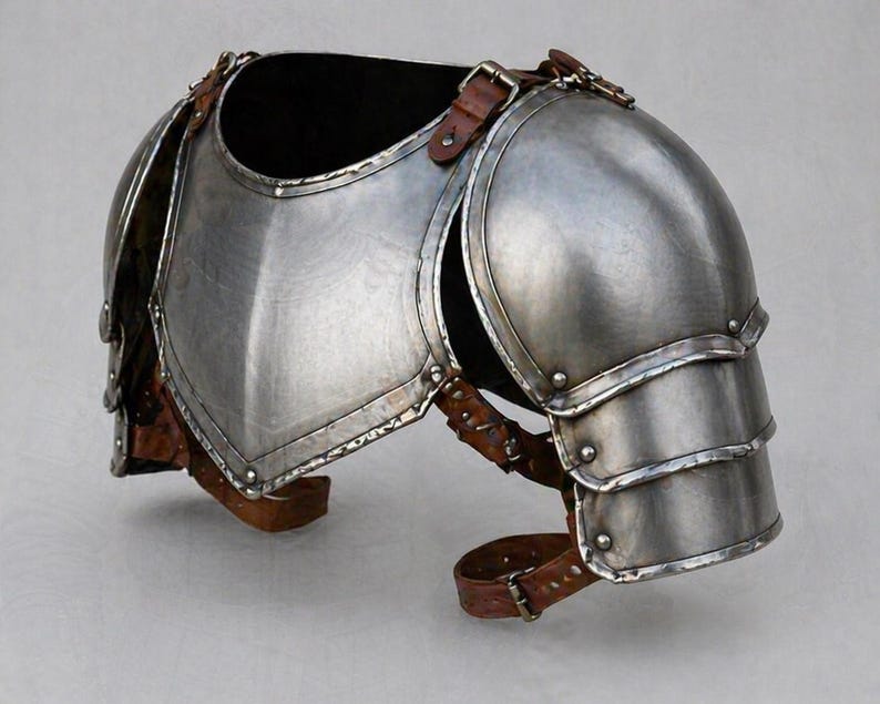 May include: A silver-colored metal shoulder armor piece with brown leather straps and buckles. The armor has a curved design to fit the shoulder and upper arm, with layered plates for protection. The leather straps are riveted to the metal.