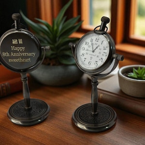 Puede incluir: Dos relojes de escritorio de estilo antiguo con marcos y soportes de metal negro. Un reloj muestra la hora, el otro tiene el texto "Happy 8th Anniversary sweetheart". Una pequeña planta en maceta y libros están en el fondo.