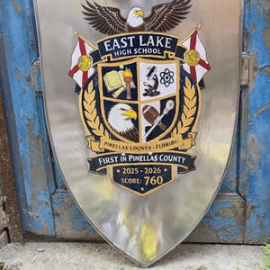 Op de afbeelding: Een zilverkleurig metalen schild met het embleem van de East Lake High School. Het wapen toont een adelaar, een boek met een fakkel, een microscoop, een atoom, een football en een lacrossecross. De tekst bevat Pinellas County, Florida, 2025-2026, Score: 760.
