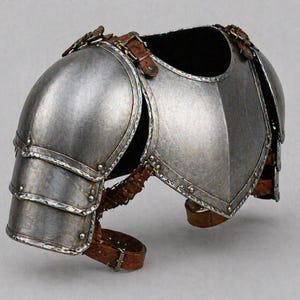 May include: A silver-colored metal shoulder armor piece with riveted construction and leather straps. The armor has a curved design to fit the shoulder and upper arm, with multiple articulated plates.