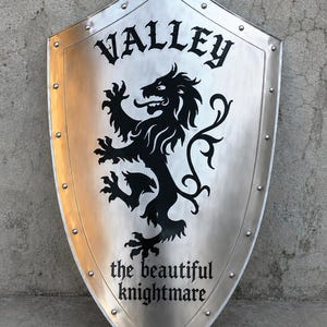 May include: A silver-colored metal shield featuring a black lion graphic in the center. The word VALLEY is printed in black above the lion, and the text the beautiful knightmare is printed in black below the lion. The shield has rivets along the edge.