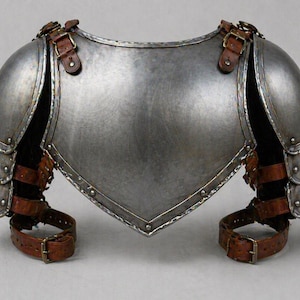 May include: A silver-colored metal chest and shoulder armor with brown leather straps and buckles. The armor has a polished finish and is designed to protect the upper body. The shoulder pieces are rounded and the chest piece is shaped to fit the torso.