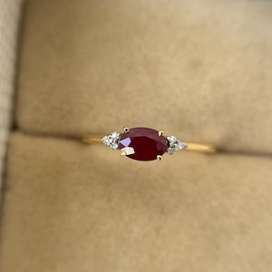Puede incluir: Un anillo de oro con un gran rubí ovalado como pieza central, flanqueado por dos piedras preciosas claras más pequeñas de forma triangular. El anillo se presenta sobre un fondo neutro.