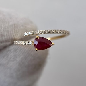 Puede incluir: Anillo de oro con una gema de rubí rojo oscuro en forma de pera. La banda del anillo está dividida, con un lado adornado con pequeños diamantes brillantes. El rubí está sujeto por garras de oro, creando un diseño elegante.