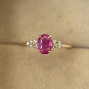 Puede incluir: Un anillo de oro con un rubí ovalado central y pequeñas piedras preciosas claras. El rubí es el punto focal, flanqueado por grupos de piedras más pequeñas. La banda del anillo es de oro pulido simple, mostrando las piedras preciosas.