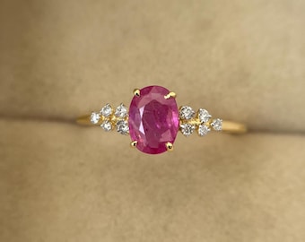 Könnte beinhalten: Ein goldener Ring mit einem ovalen Rubin im Zentrum und kleinen, klaren Edelsteinen. Der Rubin ist der Mittelpunkt, flankiert von Clustern kleinerer Steine. Das Band des Rings ist aus schlichtem, poliertem Gold, das die Edelsteine zur Geltung bringt.