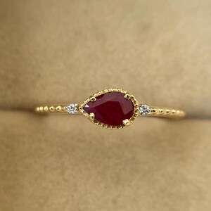 Op de afbeelding: Een gouden ring met een peervormige, dieprode robijn edelsteen. De ring heeft een delicate, kralenband en twee kleine, heldere accentstenen aan weerszijden van de robijn. De zetting rond de robijn is ook gedetailleerd met een kralenontwerp.