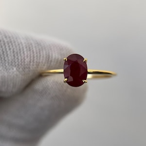May include: A gold ring featuring a large, oval-cut ruby gemstone. The ruby is a deep, rich red color and is held in place by four delicate prongs. The band of the ring is a thin, polished gold.