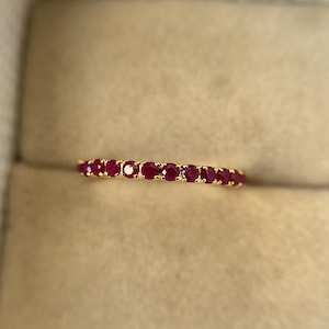 以下が含まれることがあります： 小さな鮮やかな赤い宝石が並んだゴールドバンドリング。 リングはニュートラルなベージュの背景に表示され、リングの色とデザインを強調しています。 リングはジュエリーです。