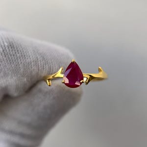 Op de afbeelding: Een gouden ring met een druppelvormige, dieprode edelsteen. De band van de ring heeft een takachtig ontwerp met kleine, puntige accenten. De edelsteen is het middelpunt, tegen een effen achtergrond.