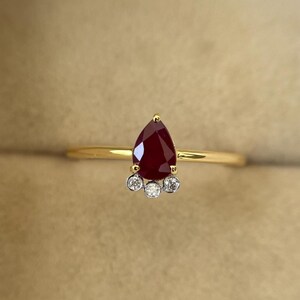 Op de afbeelding: Een gouden ring met een peervormige, dieprode edelsteen als centraal punt. Onder de rode steen bevinden zich drie kleine, ronde, heldere edelstenen. De ring heeft een dunne, gepolijste gouden band.