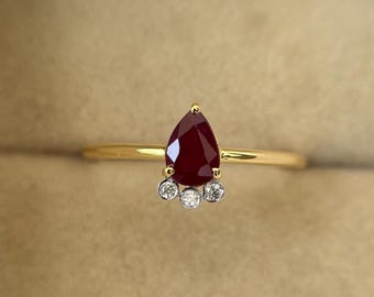 Könnte beinhalten: Ein goldener Ring mit einem tropfenförmigen, tiefroten Edelstein als Hauptmerkmal. Unterhalb des roten Steins befinden sich drei kleine, runde, klare Edelsteine. Der Ring hat ein dünnes, poliertes Goldband.