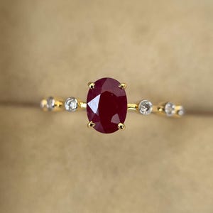 以下が含まれることがあります： 中心に楕円形の深い赤色のルビーをあしらったゴールドリング。バンドには、小さく丸い透明な宝石が飾られています。リングのデザインはシンプルでありながらエレガントで、ルビーの鮮やかな色を際立たせています。