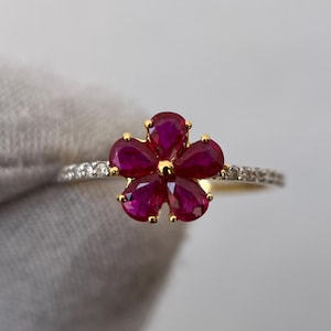 May include: A gold ring featuring a flower design with five ruby petals. The center of the flower is gold, and the band is adorned with small, clear gemstones. The ring is a piece of jewelry.