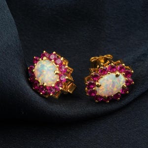 May include: A pair of gold stud earrings. Each earring has an oval, iridescent opal centre stone, encircled by small, round, pink gemstones. The earrings are set against a dark blue background.
