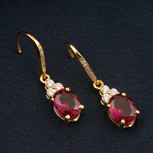 May include: A pair of gold-coloured drop earrings. Each earring features a large, oval-shaped, deep red gemstone, accented with smaller white gemstones. The earring hooks are also embellished with small, clear stones.