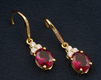 May include: A pair of gold-colored drop earrings. Each earring features a large, oval-shaped, deep red gemstone, accented with smaller white gemstones. The earring hooks are also embellished with small, clear stones.