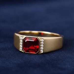 May include: A gold ring with a large, rectangular, red gemstone as the centerpiece. Small, clear stones are set on either side of the red stone. The ring is set against a dark blue background.