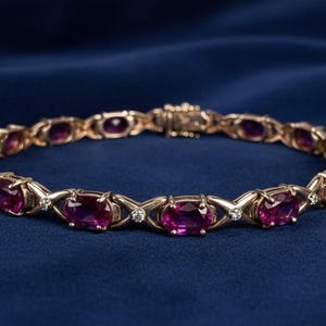 May include: Gold-toned bracelet with oval-cut, deep pink gemstones and small, round accent stones. The bracelet is set against a dark blue background, showcasing its intricate design and color.