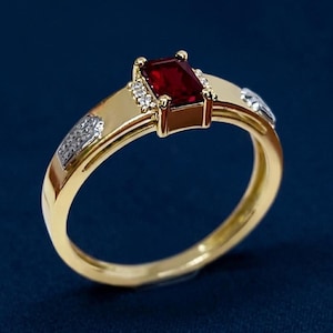 May include: A gold ring with a rectangular, deep red gemstone at the center. Small, clear stones are set on either side of the main stone. The band has a polished finish and a decorative design on the sides.