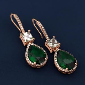 May include: A pair of rose gold drop earrings featuring a pear-shaped dark green gemstone surrounded by small clear stones. Above the pear-shaped stone is a square-cut clear gemstone. The ear wires are also accented with small clear stones.
