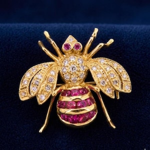 Puede incluir: Broche de abeja dorada con detalles de rubíes y diamantes. El cuerpo de la abeja presenta rayas horizontales de rubíes, mientras que las alas y el cuerpo están adornados con diamantes brillantes. Los ojos son dos pequeños rubíes.