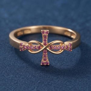 May include: A rose gold ring featuring a central cross design intertwined with an infinity symbol. The cross and the infinity loops are encrusted with small, vibrant pink gemstones that extend along the band on both sides of the centerpiece.