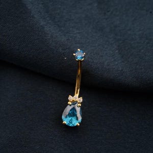 May include: A gold-coloured belly button ring featuring a teardrop-shaped, light blue gemstone. The ring has a small, round blue gem at the top and small clear gems near the teardrop. Set against a dark blue background.