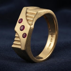 Peut inclure: Une bague en or avec une finition brossée et un design géométrique comportant trois petits rubis rouges de taille ovale disposés en diagonale. L'anneau présente une texture architecturale étagée sur les côtés et des poinçons visibles à l'intérieur.