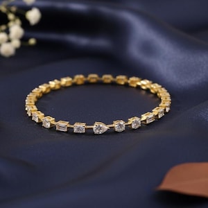 Puede incluir: Una pulsera dorada con formas alternas de piedras preciosas transparentes. La pulsera presenta piedras cuadradas, rectangulares, en forma de pera y redondas engastadas en una banda dorada. La joya se exhibe sobre un fondo azul oscuro.