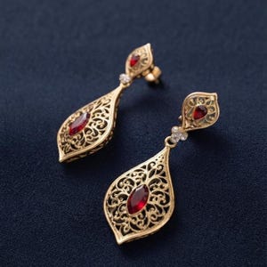 May include: A pair of gold drop earrings featuring an intricate filigree metalwork design in a teardrop shape. Each earring is centered with a marquise-cut deep red gemstone and accented with a smaller teardrop-shaped red stone at the top post.