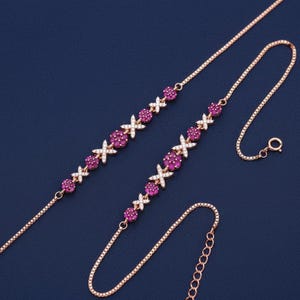 Puede incluir: Collar de oro rosa con una cadena delicada y un patrón repetido de piedras preciosas rosas y detalles blancos. El collar tiene un cierre de mosquetón y se muestra sobre un fondo azul oscuro.