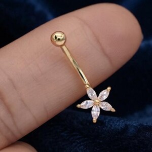 May include: A gold-toned curved barbell body jewelry piece featuring a polished spherical top bead and a flower-shaped bottom charm. The flower charm is composed of six marquise-cut clear white stones arranged around a central gold-toned bead.