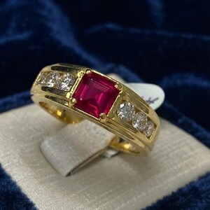 Puede incluir: Un anillo de oro con una piedra preciosa roja vibrante de corte cuadrado en el centro, flanqueada por tres piedras claras redondas a cada lado. La banda es de oro amarillo pulido y presenta un engaste de canal para las piedras.