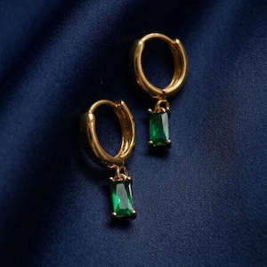 May include: A pair of gold hoop earrings, each featuring a small, rectangular, emerald green gemstone charm dangling from the bottom. The earrings have a polished metallic finish and a hinged closure mechanism, showcasing a vibrant green hue.