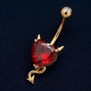May include: A gold-colored belly button ring with a red heart-shaped gemstone. The heart has small horns and a tail with an arrow. The top ball has a clear gemstone.