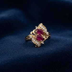 Peut inclure: Une bague en or ornée de deux rubis rouge foncé taille ovale disposés verticalement au centre, entourés d'un motif tourbillonnant serti de petits diamants blancs ronds qui s'étendent le long des épaules de l'anneau en or poli.