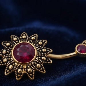 May include: A gold-toned navel ring featuring a large, sunburst-shaped charm with a central round, deep red gemstone. The curved barbell connects to a smaller, matching decorative end piece that also contains a single, smaller round red gemstone.