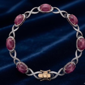May include: A circular bracelet featuring six oval cabochon star rubies set in silver-toned metal. The band consists of an intricate, twisted infinity-style link pattern encrusted with small white stones, finished with a gold-toned metal box clasp.