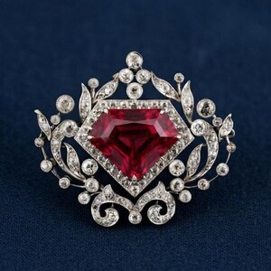 Peut inclure: Une broche argentée avec une grande pierre précieuse rouge en forme de cerf-volant au centre. La pierre est entourée d'une monture métallique décorative incrustée de nombreuses petites pierres rondes et claires en motifs de feuilles.