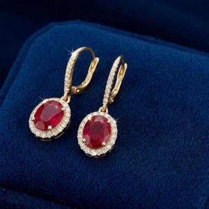 May include: A pair of gold drop earrings featuring oval-cut red gemstones surrounded by a halo of small white diamonds. The earrings have a hinged lever-back closure and a polished gold finish, showcasing a classic design with vibrant red stones.
