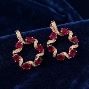 May include: A pair of circular pendants crafted from rose-toned metal, each featuring a swirling design set with multiple oval-cut deep red rubies and small white accent stones along the curved bands, creating a continuous open-center wreath shape.
