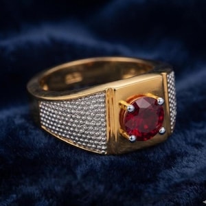 May include: A gold ring featuring a round, faceted red gemstone held by four prongs. The band is constructed with a smooth gold center section and textured, silver-toned granular side panels that extend around the circumference of the ring.