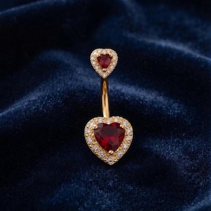 May include: A gold-toned belly button ring featuring two heart-shaped red stones. The smaller top heart and larger bottom heart are each surrounded by a halo of small, clear, sparkling stones, connected by a curved gold-toned metal bar.
