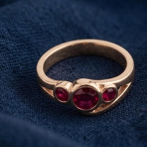 Puede incluir: Un anillo de oro con un rubí rojo central de talla redonda flanqueado por dos rubíes rojos más pequeños de talla redonda. La banda tiene un acabado liso y pulido con un diseño de vástago dividido que se estrecha hacia la montura central.