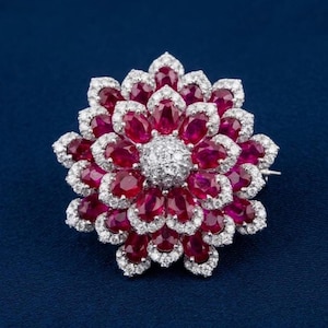 Op de afbeelding: Een bloemvormige broche met meerdere lagen ovaal geslepen dieprode robijnen. Elke robijn is omringd door een rand van kleine, heldere, fonkelende witte diamanten. Het midden van de bloem is een cluster van ronde, pavé gezette witte diamanten.