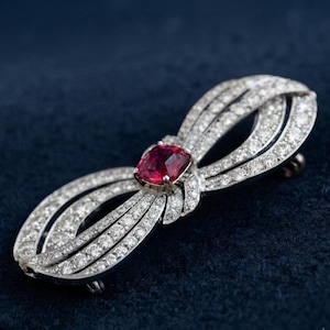 May include: A bow-shaped brooch crafted from silver-toned metal, featuring a central cushion-cut red gemstone surrounded by multiple rows of small, clear, sparkling stones set along the entire length of the looped ribbon design.