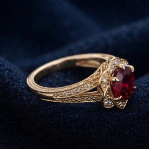 Puede incluir: Un anillo de oro con una piedra preciosa roja oscura de corte ovalado en el centro, rodeada por un halo de pequeñas piedras blancas. La banda presenta un intrincado trabajo de metal con detalles milgrain y piedras blancas.