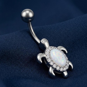 Op de afbeelding: Een zilveren navelpiercing met een schildpadontwerp. De schildpad heeft een witte opaal middensteen omringd door heldere kristallen. De gebogen barbell heeft een zilveren bal aan de bovenkant.